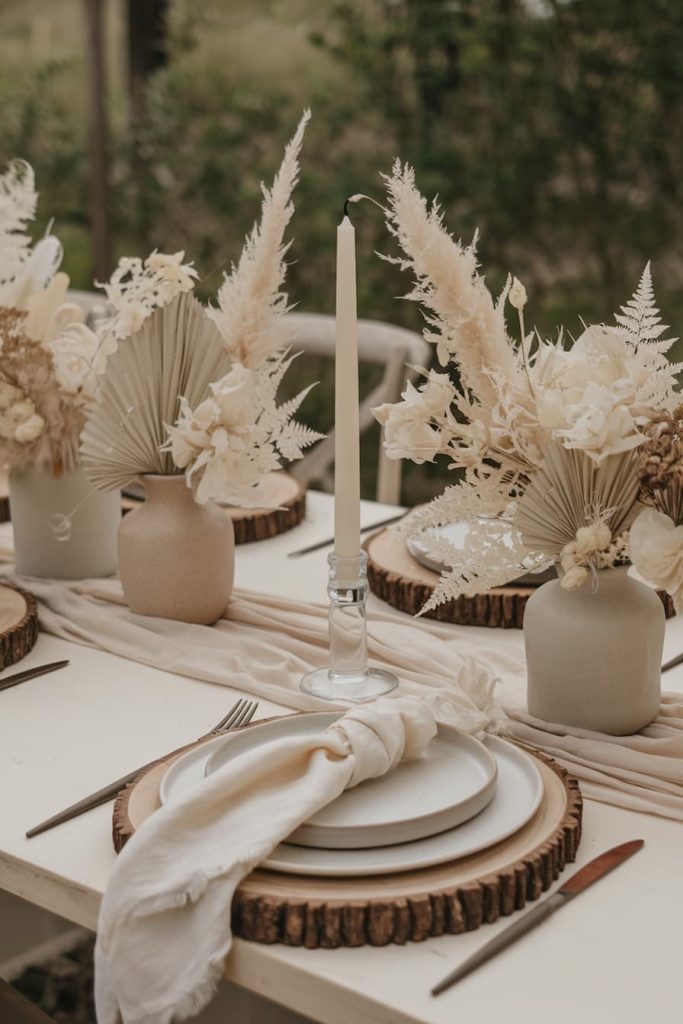 A rustic table with white plates on wooden plates, beige napkins, tall white candles and vases full of <a href=