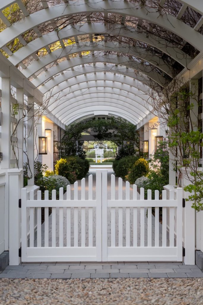 White picket fence gate under a pergola with climbing plants leading to a garden path lined with greenery and lit by wall lanterns.