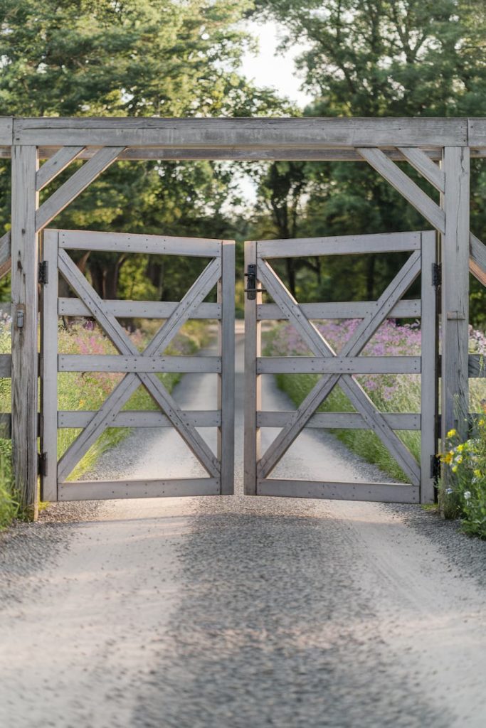 A dirt road passes through an open wooden gate with a cross pattern surrounded by green trees and wildflowers on a sunny day.