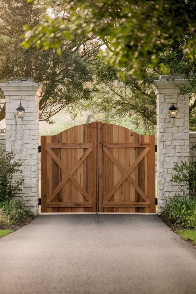 A wooden double gate closed off a paved driveway, flanked by stone pillars with lanterns and surrounded by lush greenery.