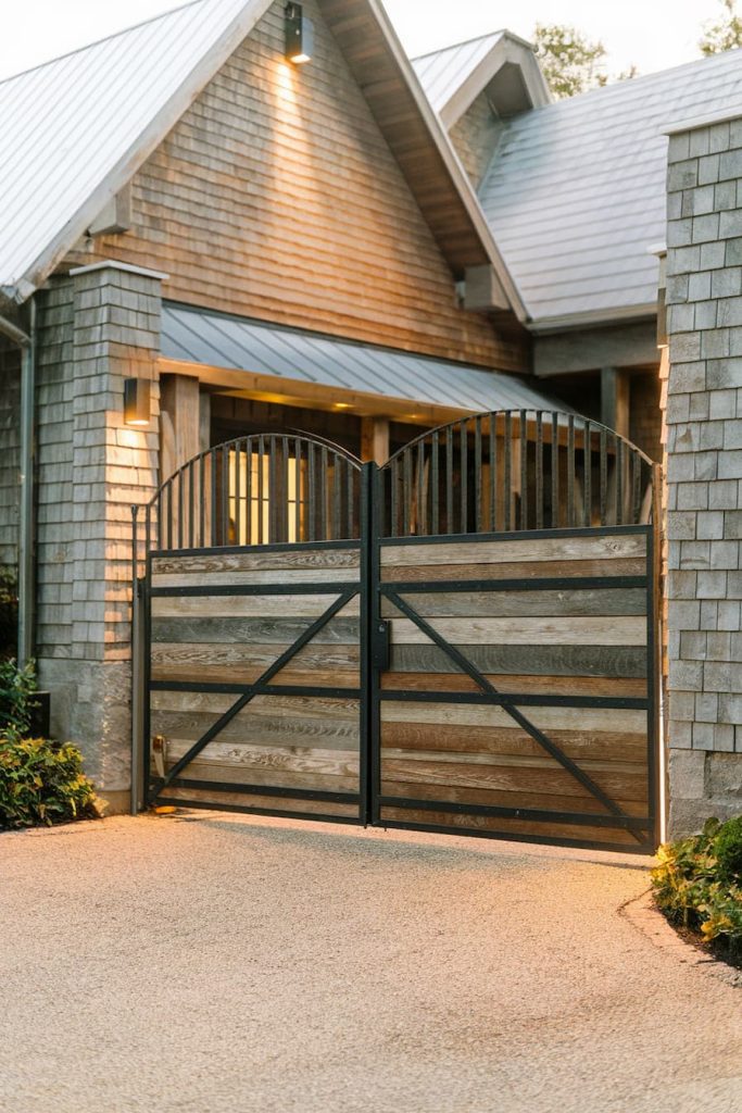 A wooden and metal gate in front of a shingle-style house with a peaked roof surrounded by stone pillars.