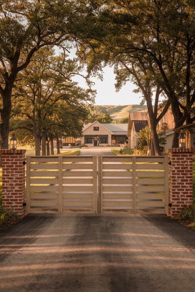 A rustic farmhouse is visible through a wooden gate flanked by brick pillars, and a tree-lined path leads to the house.