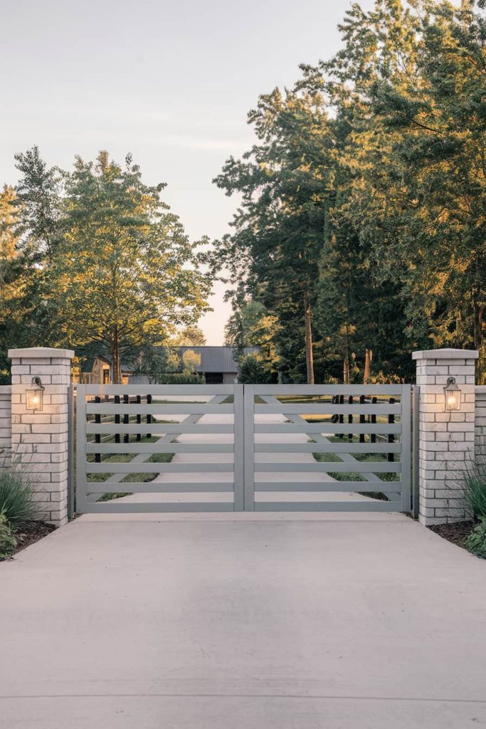 A modern wooden gate with horizontal slats leads to a driveway flanked by brick pillars and surrounded by trees under clear skies.