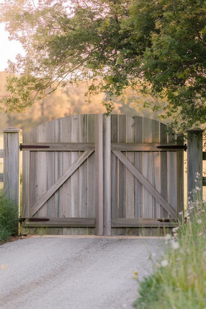 A wooden gate is closed opposite a driveway, surrounded by greenery and trees, and soft natural light illuminates the scene.