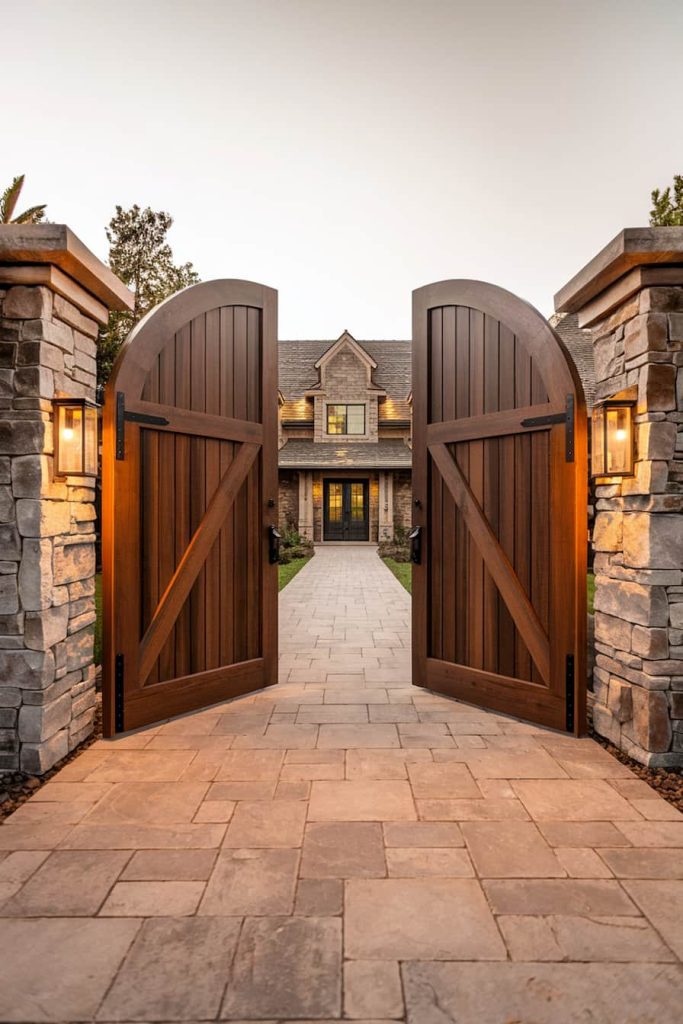 Large wooden gates open to reveal a stone walkway leading to a magnificent house surrounded by stone pillars with lanterns.