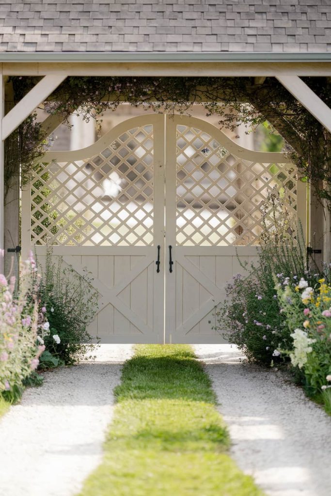 A white lattice garden gate under a covered arch surrounded by various blooming flowers opens onto a gravel and grass path.