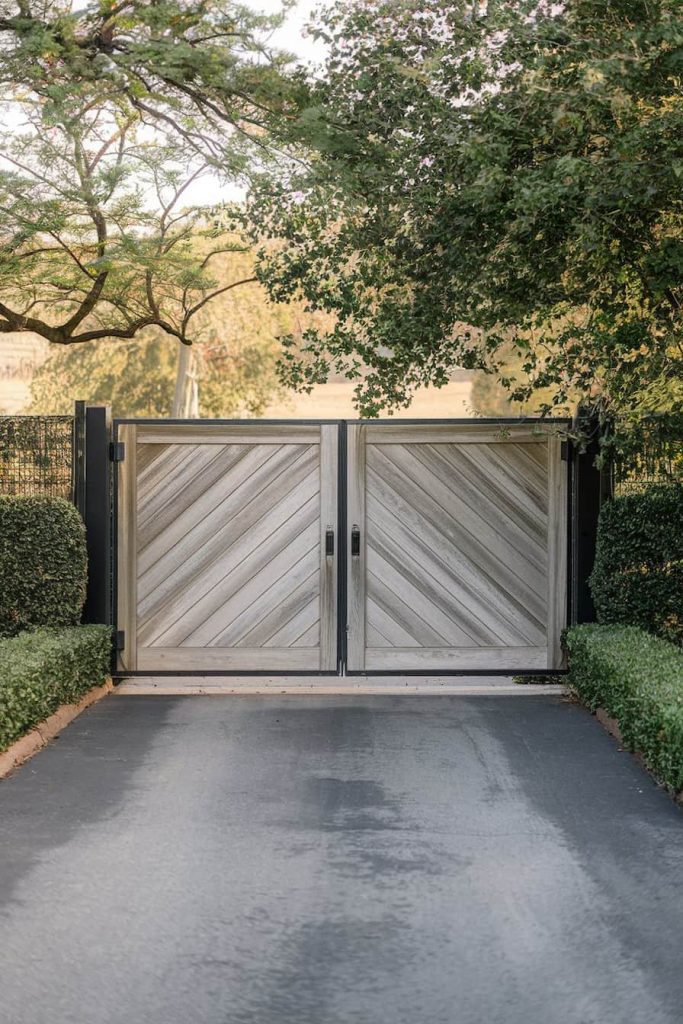 A closed wooden gate with diagonal panels surrounded by greenery and trees on a paved driveway.