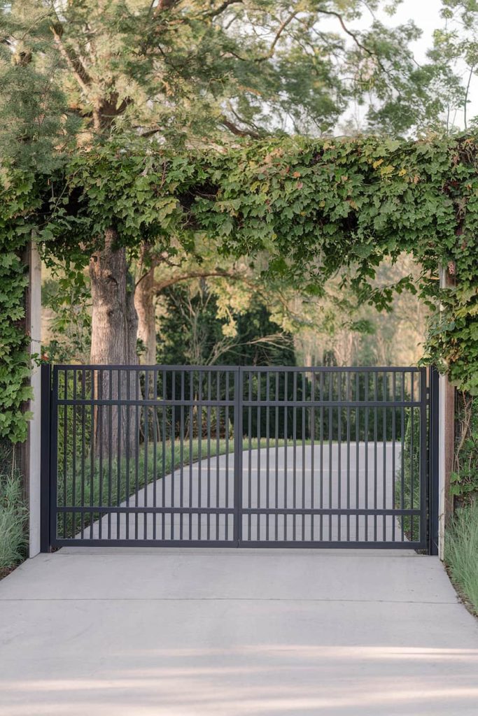 A black metal gate with vertical bars stands at the entrance to a driveway surrounded by green vines and trees.