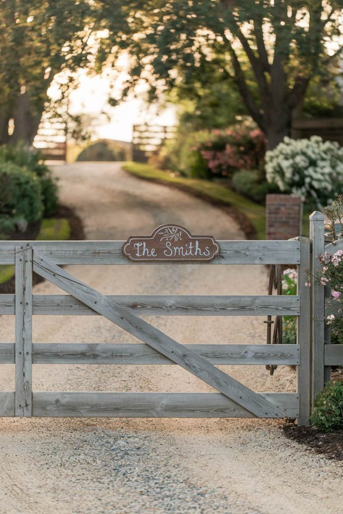 Wooden gate with a sign saying "The Smiths" in a rural setting leading to a gravel driveway lined with trees and flowers.