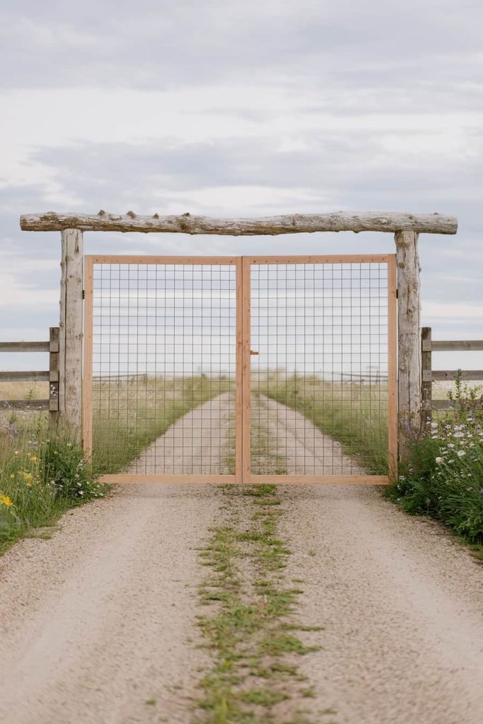 A wooden gate with wire mesh stands at the entrance to a gravel road flanked by wooden fences. Cloudy sky in the background.