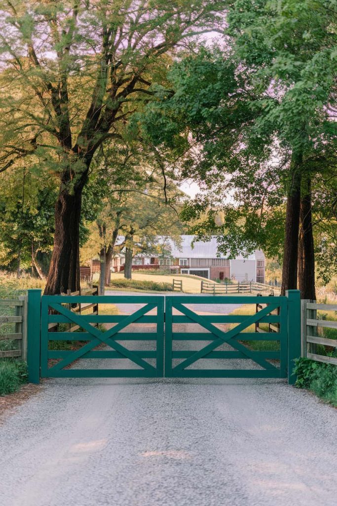 A gravel path leads to a green wooden gate flanked by tall trees, with a farmhouse and barn visible in the distance.