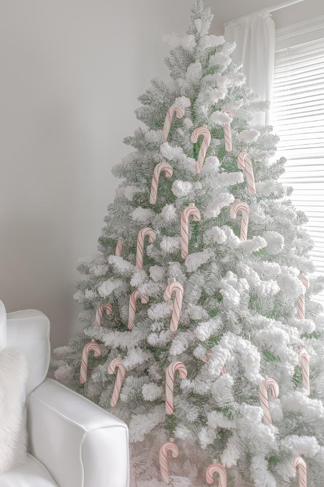 A white Christmas tree decorated with candy canes stands next to a white armchair in a brightly lit room.