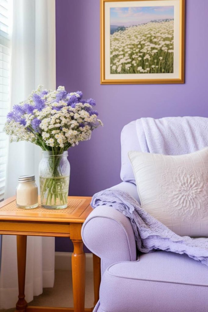 A cozy lavender-themed room features a plush armchair, a side table with a candle and a vase of white and purple flowers, and a framed floral field painting on the wall.