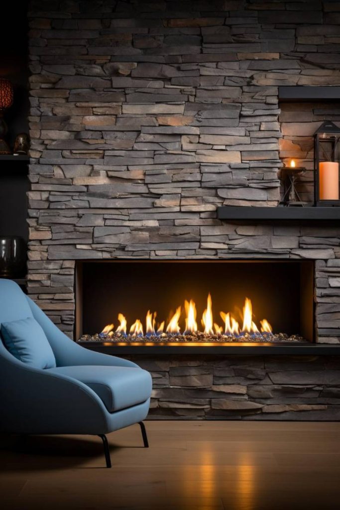 A blue chair stands by a modern stone fireplace with burning flames, next to shelves with a lantern and candles.