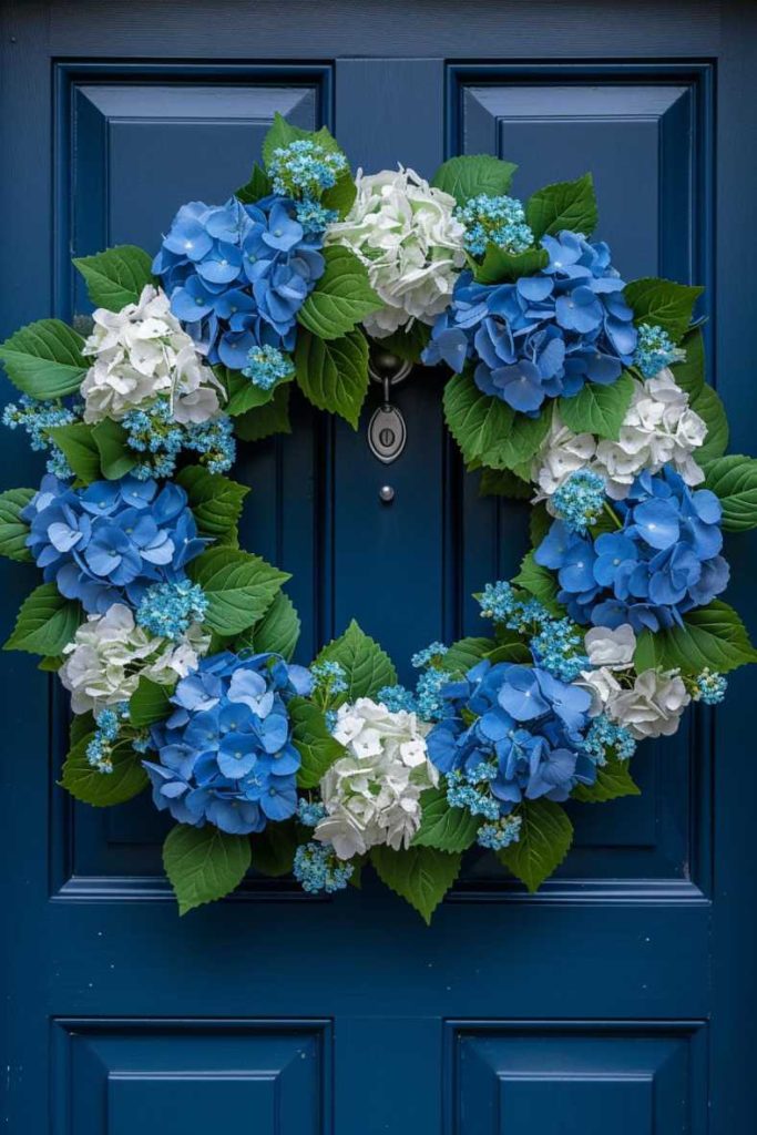 A blue door hangs in the center with a wreath of blue and white hydrangeas accented by green leaves and small blue flowers.