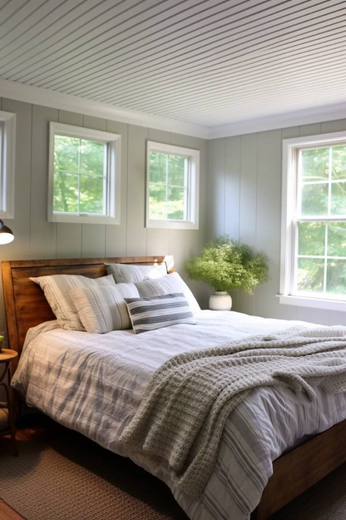 A cozy bedroom with a soft bed decorated with striped linens and a knitted throw. Four windows line the white-paneled walls, and a potted plant sits on a nightstand, bathed in natural light.