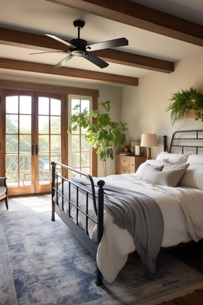 A bedroom with a black metal bed, a white bedspread, a gray blanket, wooden ceiling beams, a ceiling fan, a bedside table with a lamp, a potted plant and large windows with a view of the greenery.