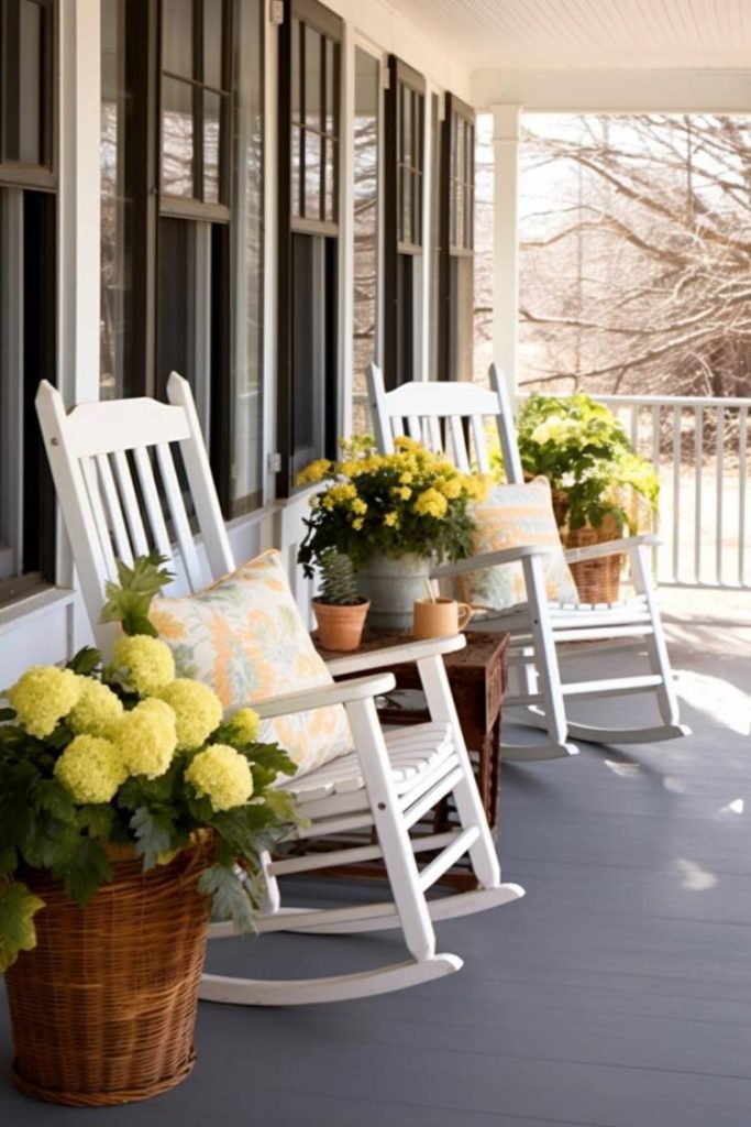 A front porch with white rocking chairs, yellow floral arrangements in wicker baskets, decorative pillows and potted plants, next to a wooden railing.