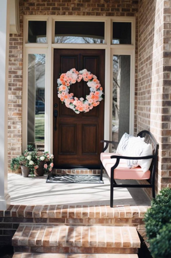 A wooden front door with a floral wreath flanked by sidelights. On the porch there is a bench with cushions accompanied by potted plants.