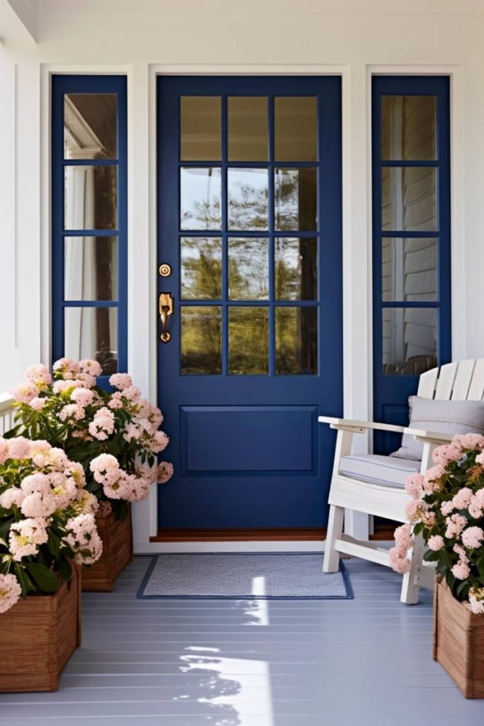 A blue glass-paned front door flanked by sidelights is framed by potted pink hydrangeas and a white wooden chair on a gray porch.