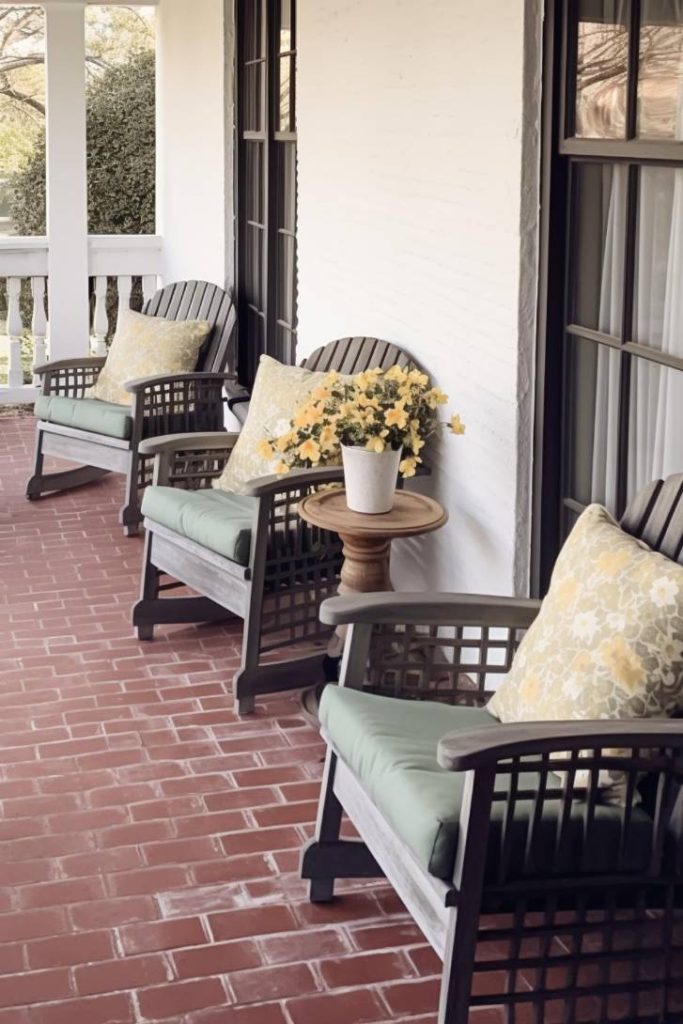 Three wooden chairs with cushions on a brick porch accompanied by a small round table with a white pot of yellow flowers.