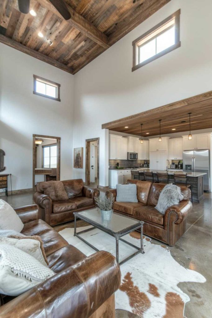 A spacious living room with high ceilings, brown leather sofas, a glass coffee table and a cowhide rug. The space leads into an open kitchen with white cabinets and stainless steel appliances.