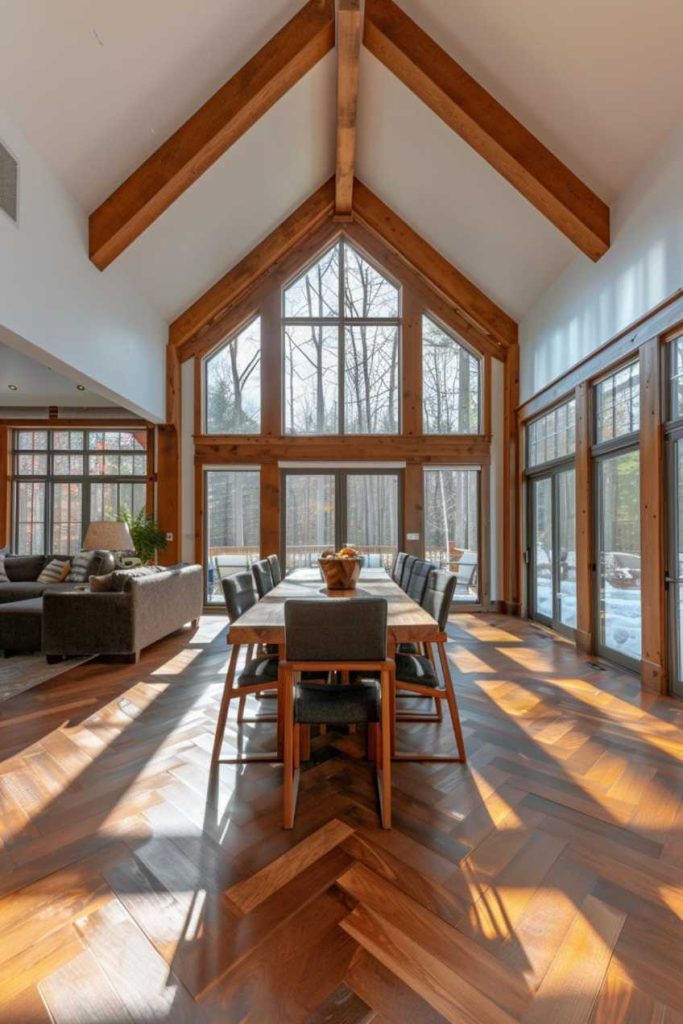 A spacious dining room with a wooden table and chairs, large windows, high wooden beams and views of the trees outside.