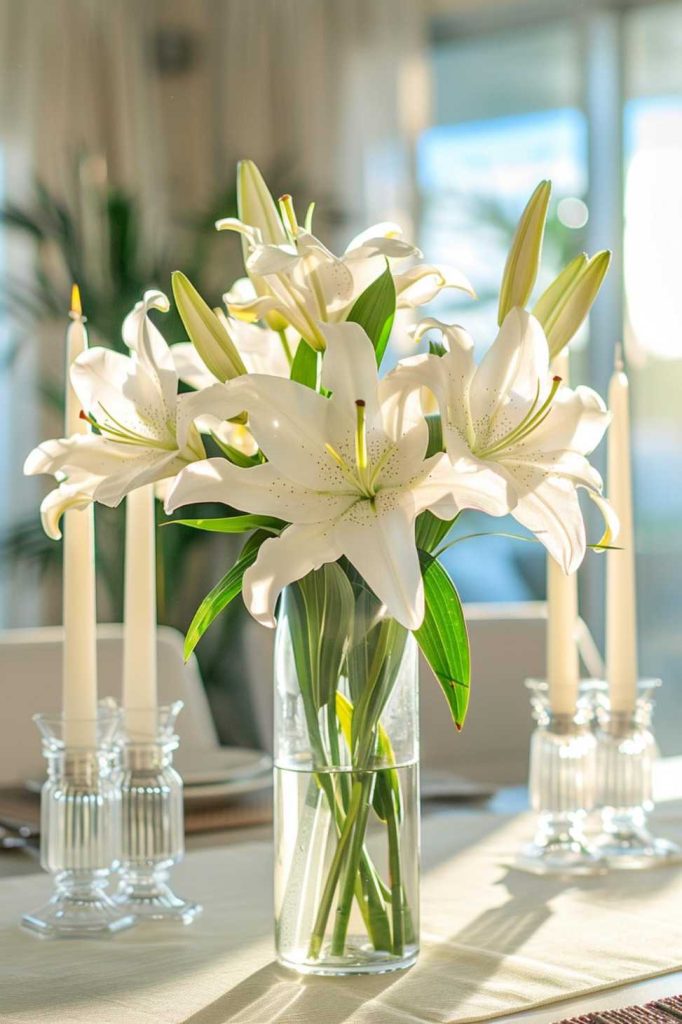 A clear glass vase of white lilies sits on a table set for dinner, flanked by two lit candles in <a href=