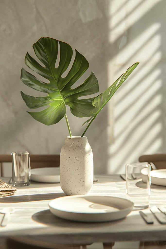 A white-flecked vase with two large green leaves sits on a sunlit dining table with white plates, glasses and cutlery.