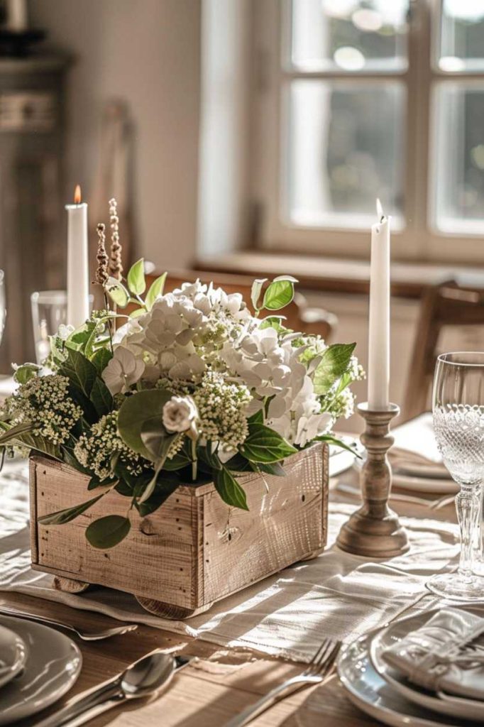 A wooden box filled with white flowers and greenery serves as the centerpiece on a table set for a meal with lit candles and glassware.