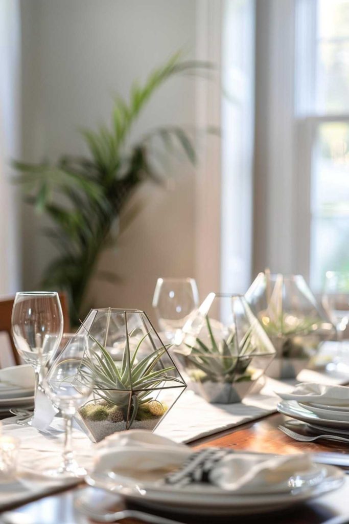 A dining table with white plates, glasses and decorative glass terrariums with small plants; A large plant can be seen in the background next to the windows.