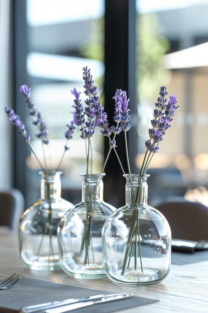 Three clear glass vases with purple lavender sprigs stand on a table with cutlery nearby in a bright, modern room.