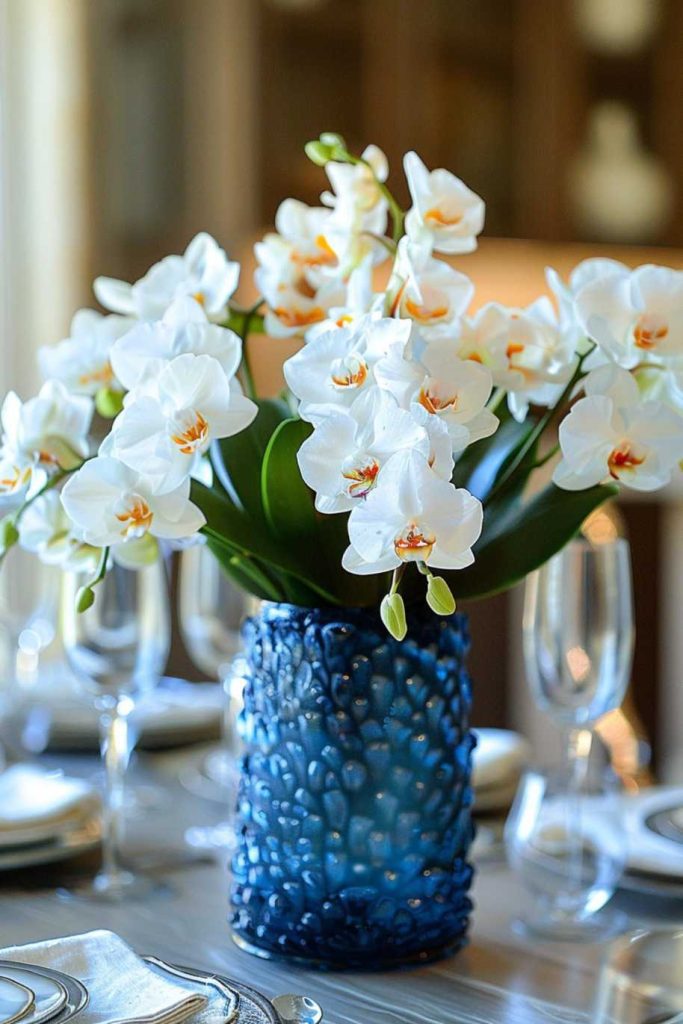 White orchids in a textured blue vase take center stage on a dining table with glassware and white plates.