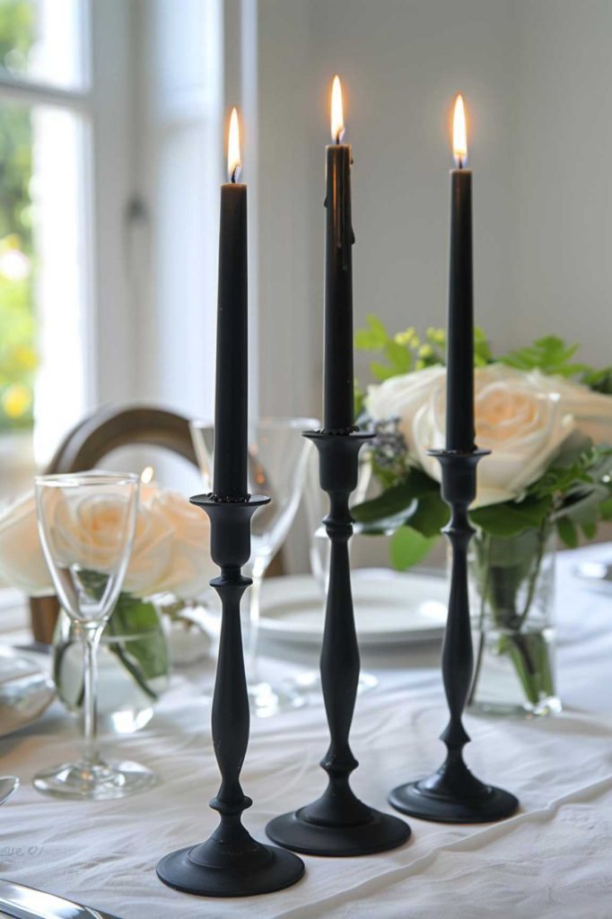 Three burning black candlesticks on a dining table with a white tablecloth surrounded by glassware and white roses in the background.