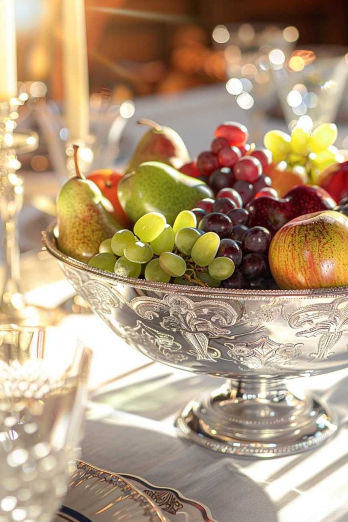 On a table set for dinner there is a silver bowl with various fruits, including grapes, apples and pears.