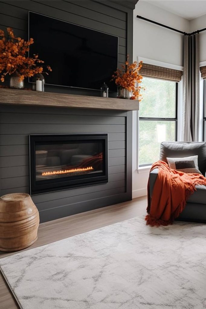 Modern living room with a dark fireplace, a lighted built-in gas fireplace, an above-mounted television, and orange fall decor. Cozy gray chair with orange blanket and large windows providing natural light.