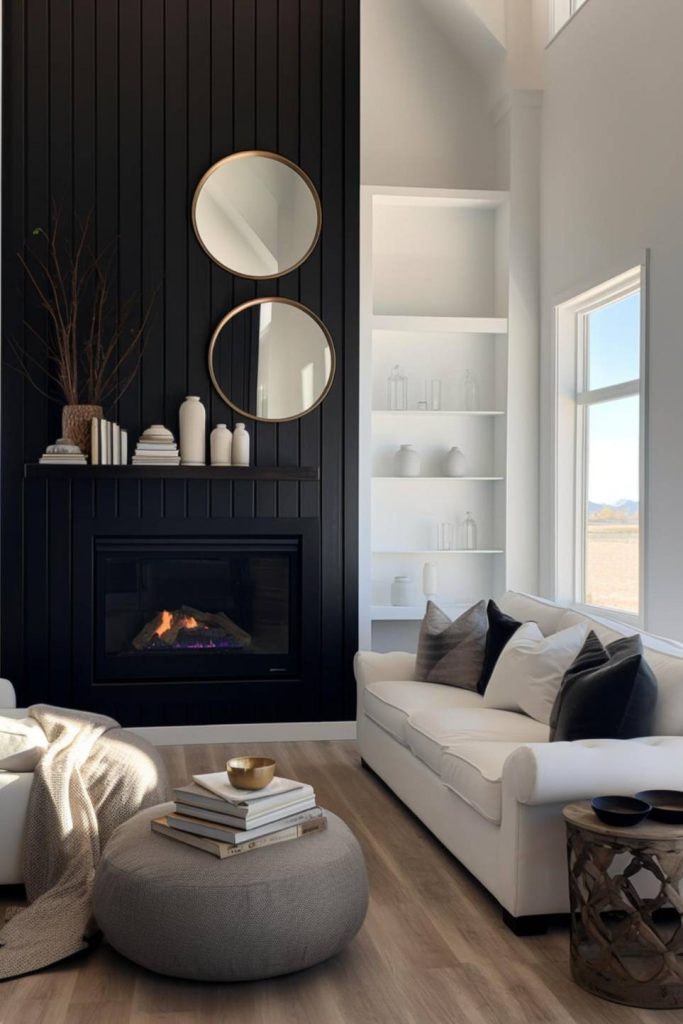 A modern living room features a black shiplap fireplace wall with two round mirrors, white shelves, a white sofa with black pillows, a gray stool and a window with a scenic view.