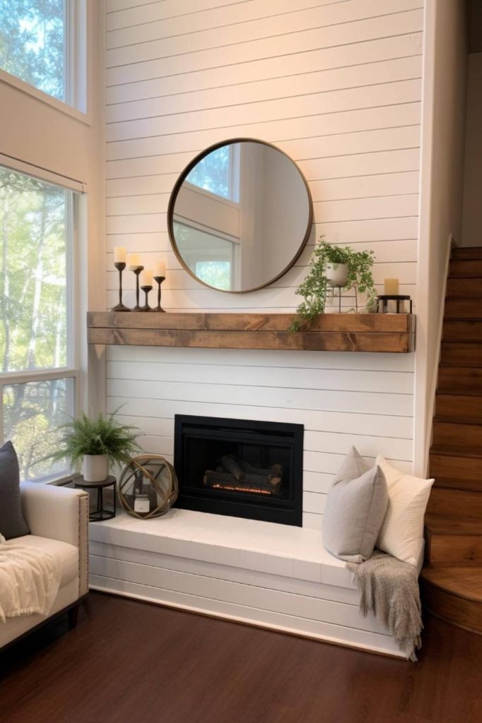 A modern living room with a gas fireplace, a round mirror, candlesticks and potted plants on a wooden mantel, white shiplap walls, large windows, a gray armchair and a wooden staircase.