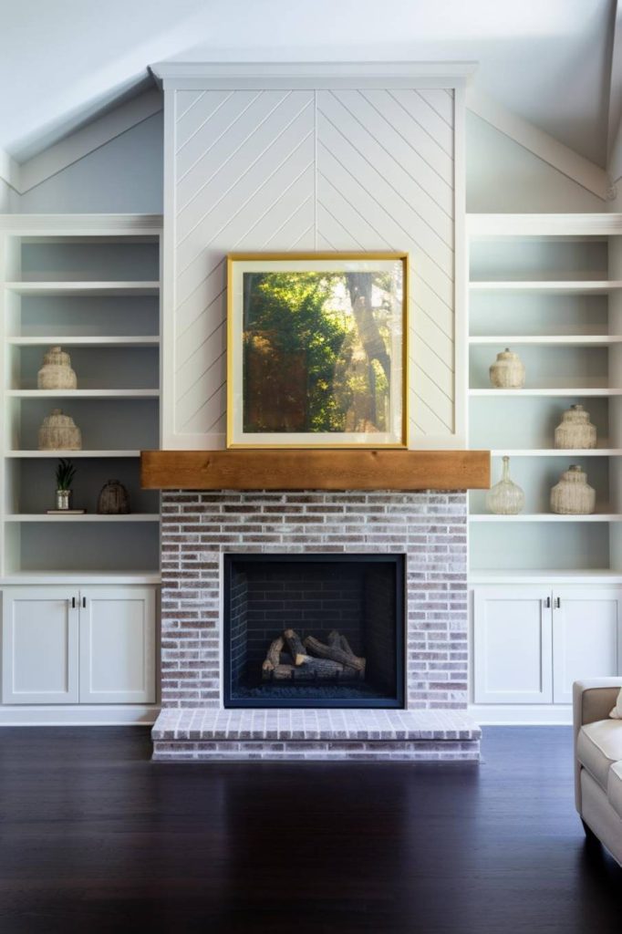 A modern living room features a fireplace with a framed picture above it, surrounded by built-in shelves with decorative items against a background of white and light gray tones.