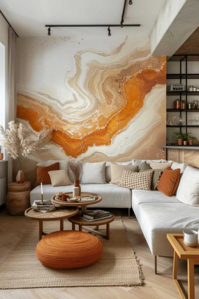 A modern living room features a white sectional sofa, nested coffee tables, a round orange stool, pampas grass in a vase, and an abstract orange and white wall mural. In the background there are shelves with decor.