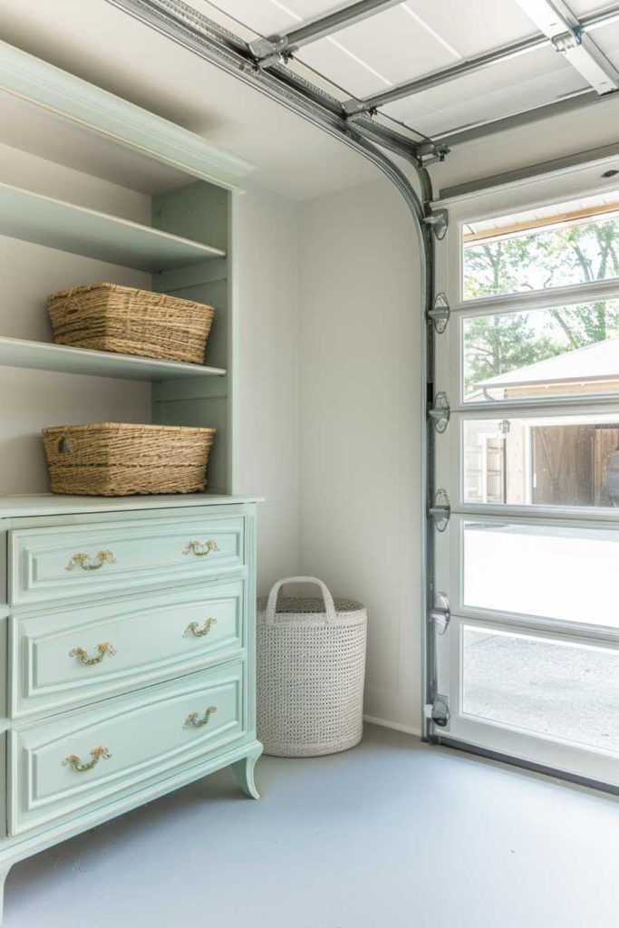 A bright garage interior with an open door, a mint green dresser, two wicker baskets on a shelf and a white laundry basket next to it.