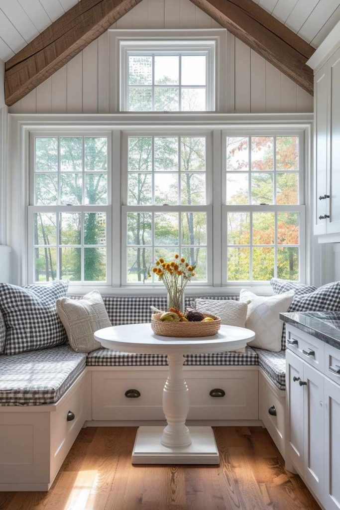 A cozy kitchen nook with a round white table, seating with gingham cushions and a window view of trees. On the table there is a flower arrangement in a vase and a basket of lemons.