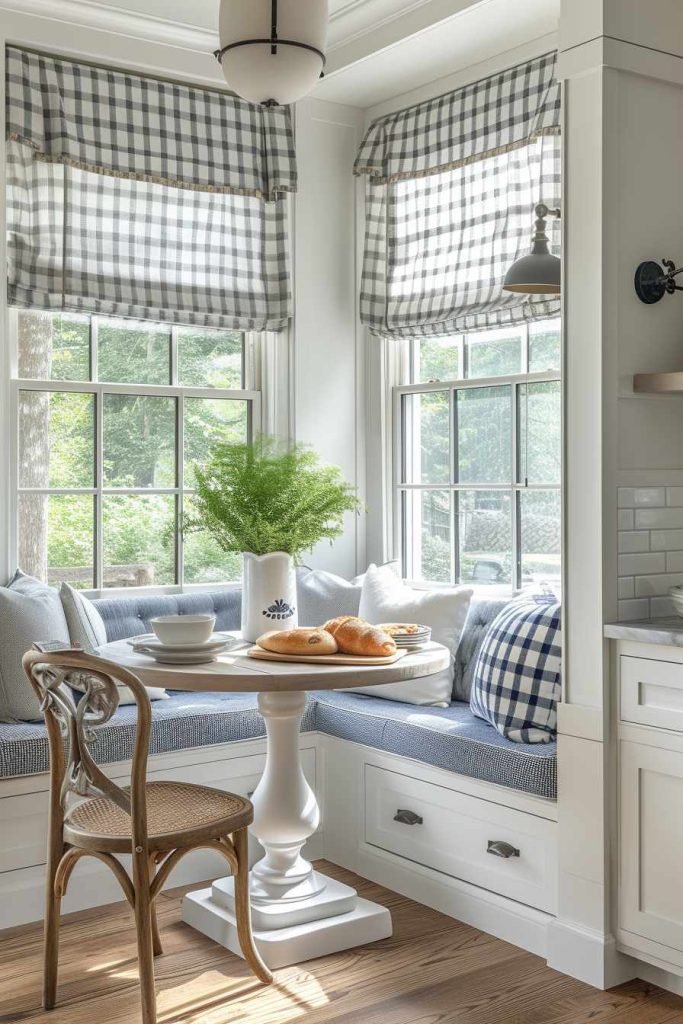 A cozy breakfast nook with a round table, wooden chair and cushioned seating next to windows with checkered curtains. On the table there is a vase with green plants, a bowl and a plate with pastries.