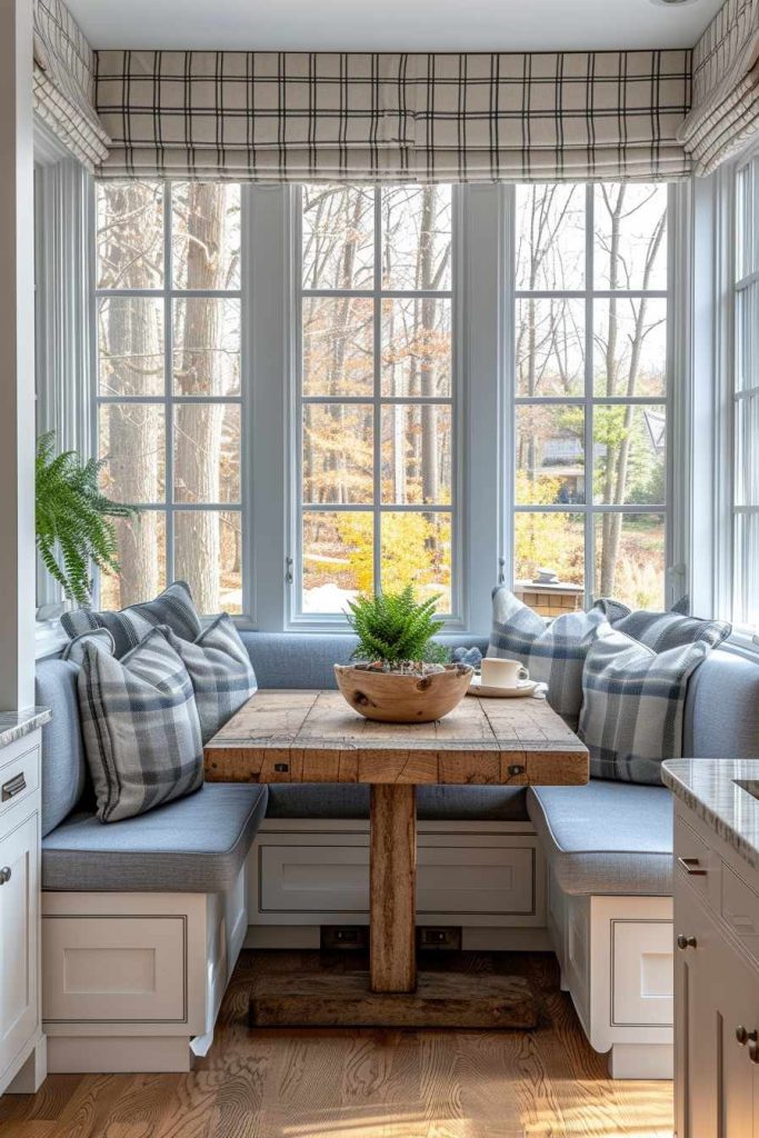 A cozy breakfast nook with a wooden table, upholstered bench seating, checkered cushions, a bowl of greenery and large windows overlooking the trees outside.