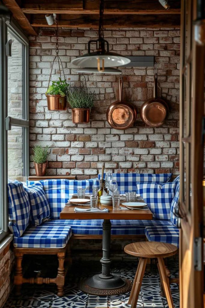 A cozy restaurant corner with a wooden table, blue and white checkered seating, brick walls, hanging copper pots, plants and set eating cutlery by the window.