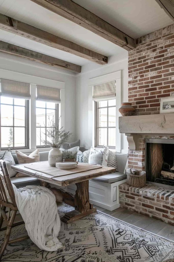 A cozy dining area with a wooden table, upholstered bench and rustic chairs. On the right side there is a brick fireplace and on the left side of the room there are windows that let in light.