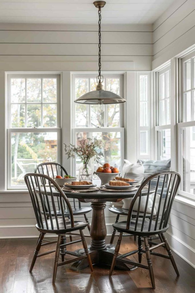 A cozy dining area with a round table, four wooden chairs, a metal pendant light and a display of fruits and flowers in front of large windows with white trim.