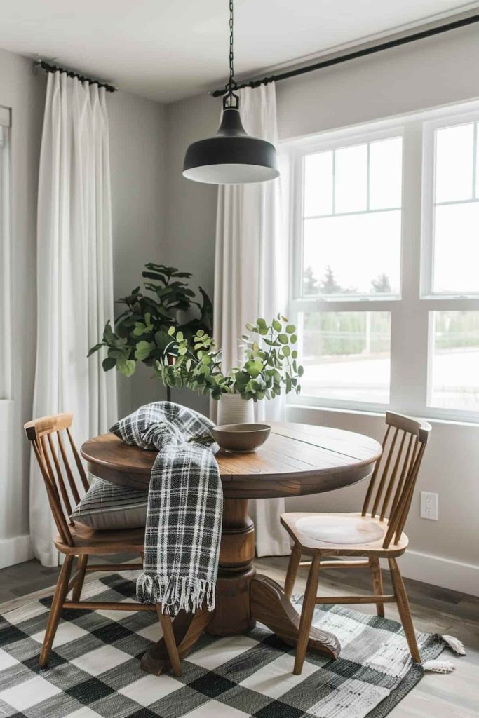 There is a small, round wooden dining table with two chairs in front of a window. There is a potted plant and a checkered blanket on the table. A black pendant light hangs above, and there is a checkered carpet underneath.