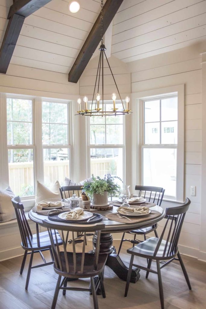 Next to large windows there is a round dining table with place settings for four people. The table features a centerpiece with a potted plant. The room features light walls and a modern chandelier.