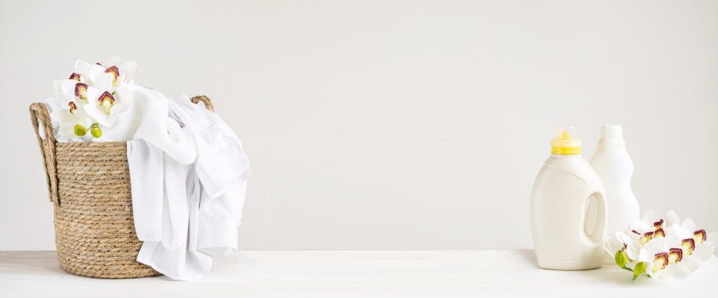 Wicker basket with white linen, washing gel and fabric softener on a white table with orchid flowers. Mockup header laundry day with copy space.